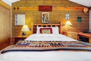 A log cabin-style bedroom with a bed, two bedside lamps, a nightstand, a dresser, and a wooden wall with framed pictures. - Country townhome next to golf course with mountain view & vaulted ceilings (Jackson)