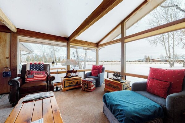 Une pièce chaleureuse avec une grande fenêtre, un plafond en bois, un fauteuil en cuir, un canapé, une table basse en bois et une petite table ornée d’objets décoratifs.