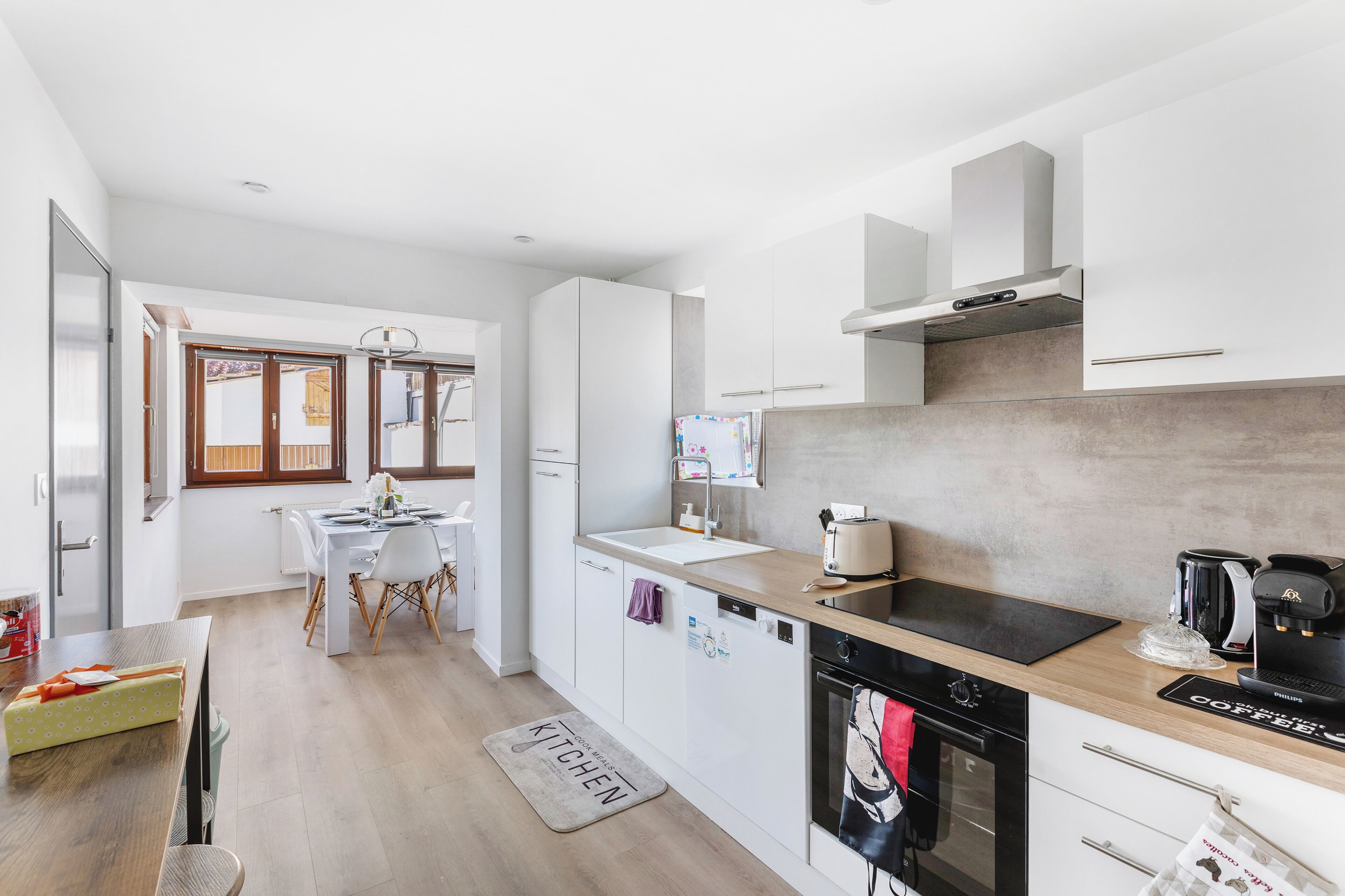 Een moderne keuken met witte kasten, een inbouwoven, een koffiezetapparaat en een eethoek met een tafel voor twee personen.