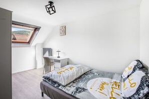 Un dormitorio con cama, escritorio, silla, radiador y ventana con vista a un tejado.