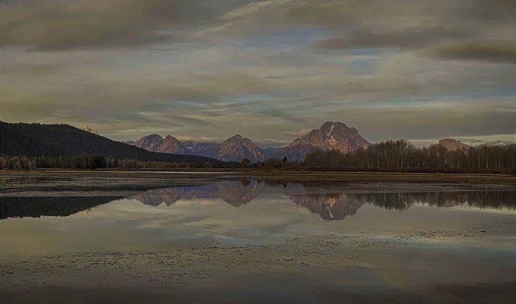 Ein ruhiger See, der Berge und einen bewaldeten Hang spiegelt.