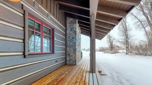 Una terraza de madera con un pilar de piedra, un ventanal de marco rojo y un paisaje cubierto de nieve.