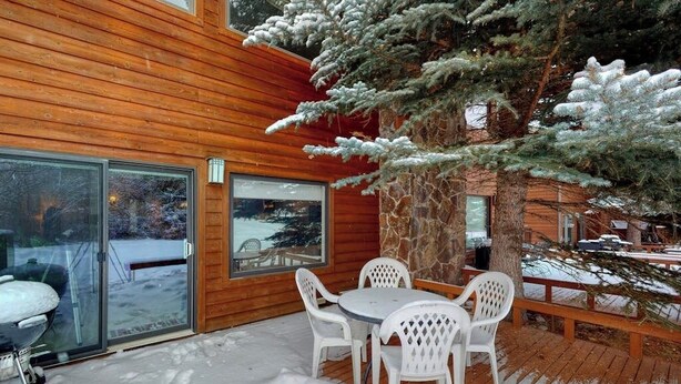 A wooden cabin with a glass door leading to a snowy deck, a round table with chairs, and a large evergreen tree. - Welcoming, well-equipped condo with furnished deck, jetted tub & fireplace (Jackson)