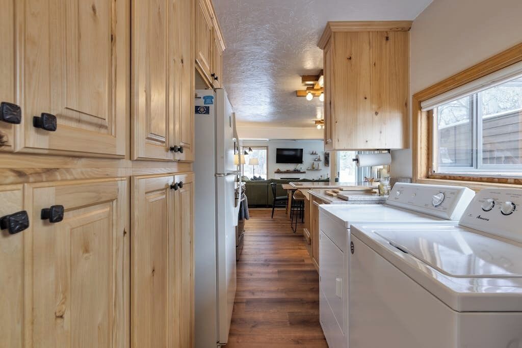 Un área de lavandería con armarios de madera, una lavadora y una zona de cocina con ventana.