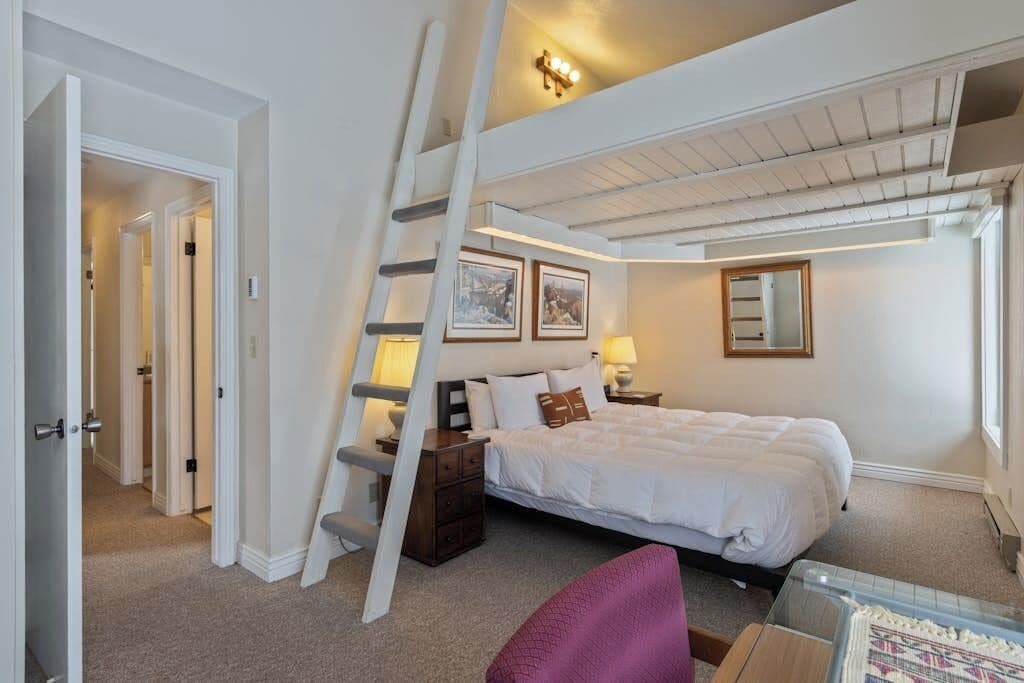 A bedroom with a loft bed, a ladder, a nightstand, a chair, and a window.