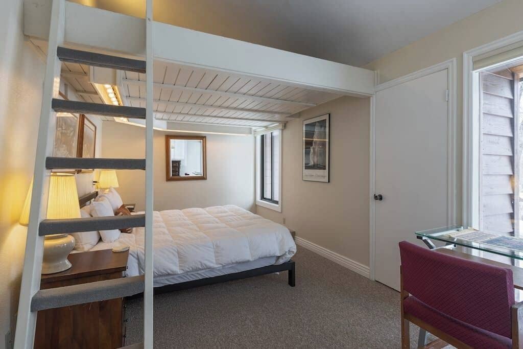 A bedroom with a bed, a desk, a chair, a ladder, and a window.