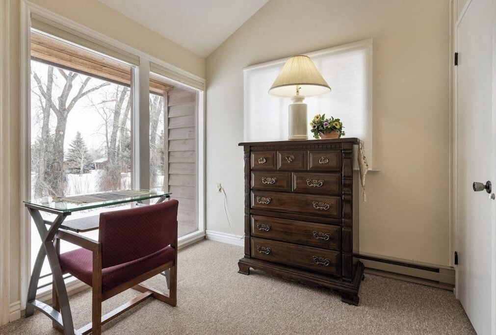 A room with a glass table, a red chair, a wooden dresser, and a lamp.
