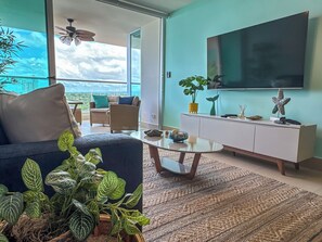 A modern living room with a sofa, coffee table, and a TV on a white cabinet. There is a balcony with a view of the sky and clouds. - Stunning Beachfront Condo in P.H. Bahia (Nueva Gorgona)