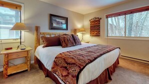 A bedroom with a bed, bedside lamps, a wooden nightstand, a framed picture on the wall, and a window with a view of trees.