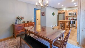 Un comedor con mesa de vidrio, sillas de madera, candelabro y cocina con armarios y electrodomésticos de madera.