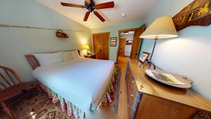 Un dormitorio ordenado con una cama, una mesita de noche de madera, un escritorio con una lámpara, un ventilador de techo y una puerta que da a otra habitación.