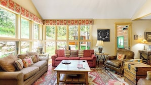 Un salon chaleureux avec un canapé, un fauteuil, une table basse et un tapis à motifs. De grandes fenêtres laissent entrer la lumière naturelle et offrent une vue sur la verdure environnante.