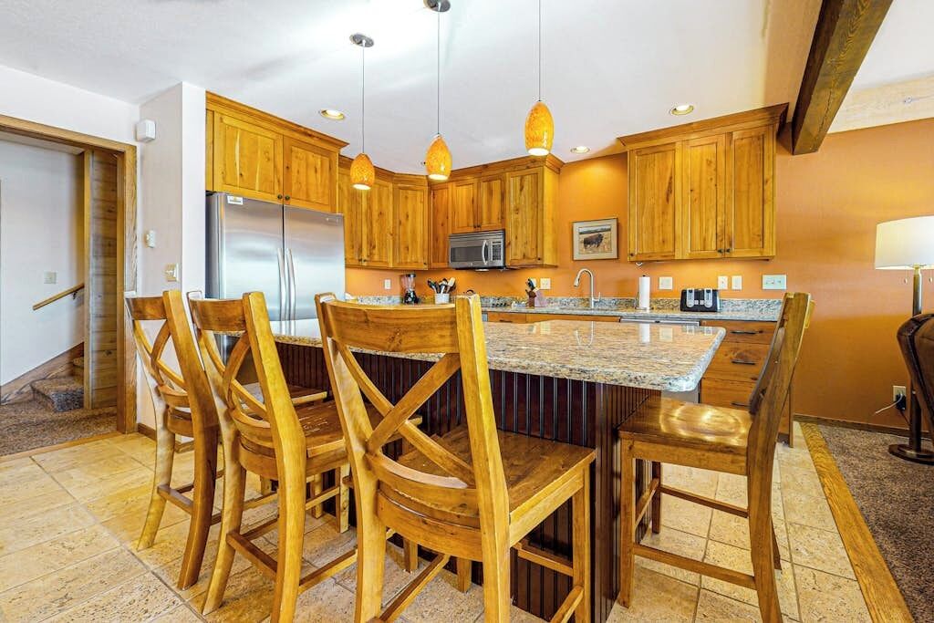 A kitchen with wooden cabinets, a stainless steel refrigerator, a microwave, and a granite countertop island with four wooden chairs.