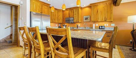 A kitchen with wooden cabinets, a stainless steel refrigerator, a microwave, and a granite countertop island with four wooden chairs.