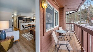 Una terraza de madera con mobiliario exterior, una cocina visible a través de un ventanal, y una vista de un paisaje nevado.