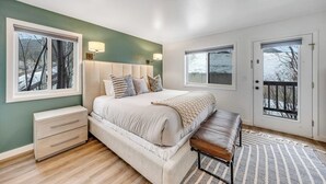 Un dormitorio con una cama grande, mesitas de noche, un banco y dos ventanas.