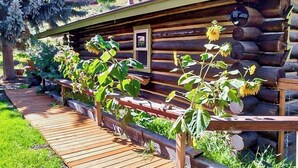 A log cabin with a wooden deck and a garden with sunflowers. - Dog-friendly waterfront cabin on Flat Creek with wood stove, creek view, firepit (Jackson)