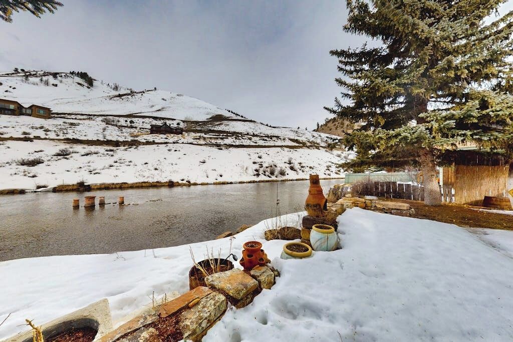 A snowy landscape with a river, a wooden structure, and a coniferous tree.