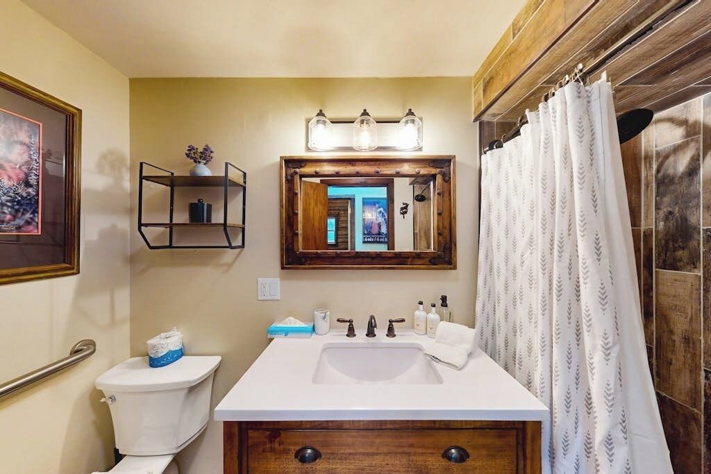 A bathroom with a white sink, wooden vanity, a mirror with lights, a toilet, and a shower curtain.