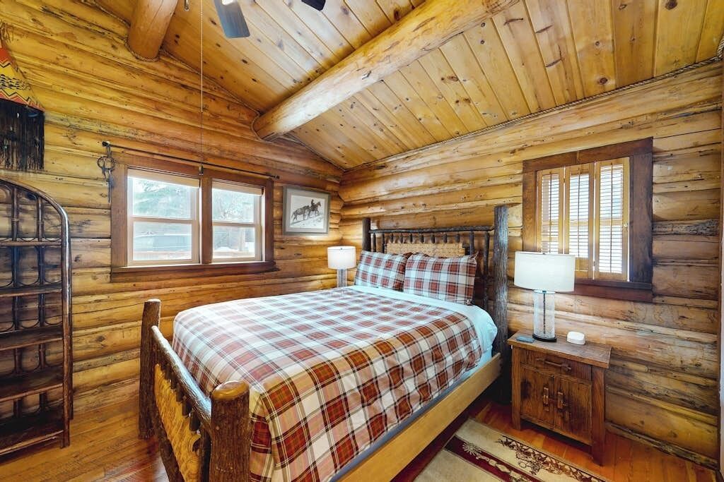 A log cabin bedroom with a bed, a nightstand, a lamp, a window, and a picture on the wall.