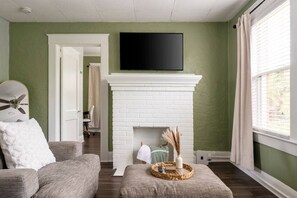 Una sala de estar con chimenea de ladrillo blanco, un televisor de pantalla plana montado encima y un sofá, además de una ventana con cortinas.