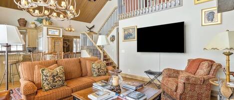 A spacious living room with a high ceiling, a large chandelier, a fireplace, a television, and a staircase.