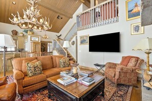 A spacious living room with a high ceiling, a large chandelier, a fireplace, a television, and a staircase. - Impressive mtn-view home near national parks with deck, fireplace, & streaming (Jackson)