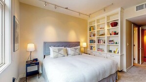Ein ordentlich eingerichtetes Schlafzimmer mit einem großen Bett, Nachttischen, einem eingebauten Bücherregal und einer Tür zu einem anderen Raum.