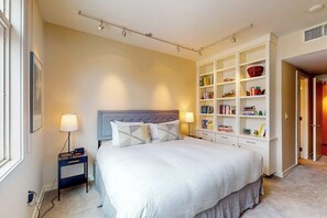 Ein ordentlich eingerichtetes Schlafzimmer mit einem großen Bett, Nachttischen, einem eingebauten Bücherregal und einer Tür zu einem anderen Raum.