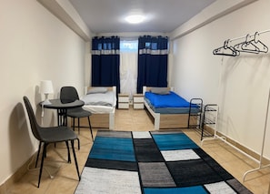Ein kleiner, gut beleuchteter Raum mit einem Bett, zwei Nachttischen, einem Sessel, einem Schreibtisch und einer Kleiderstange.