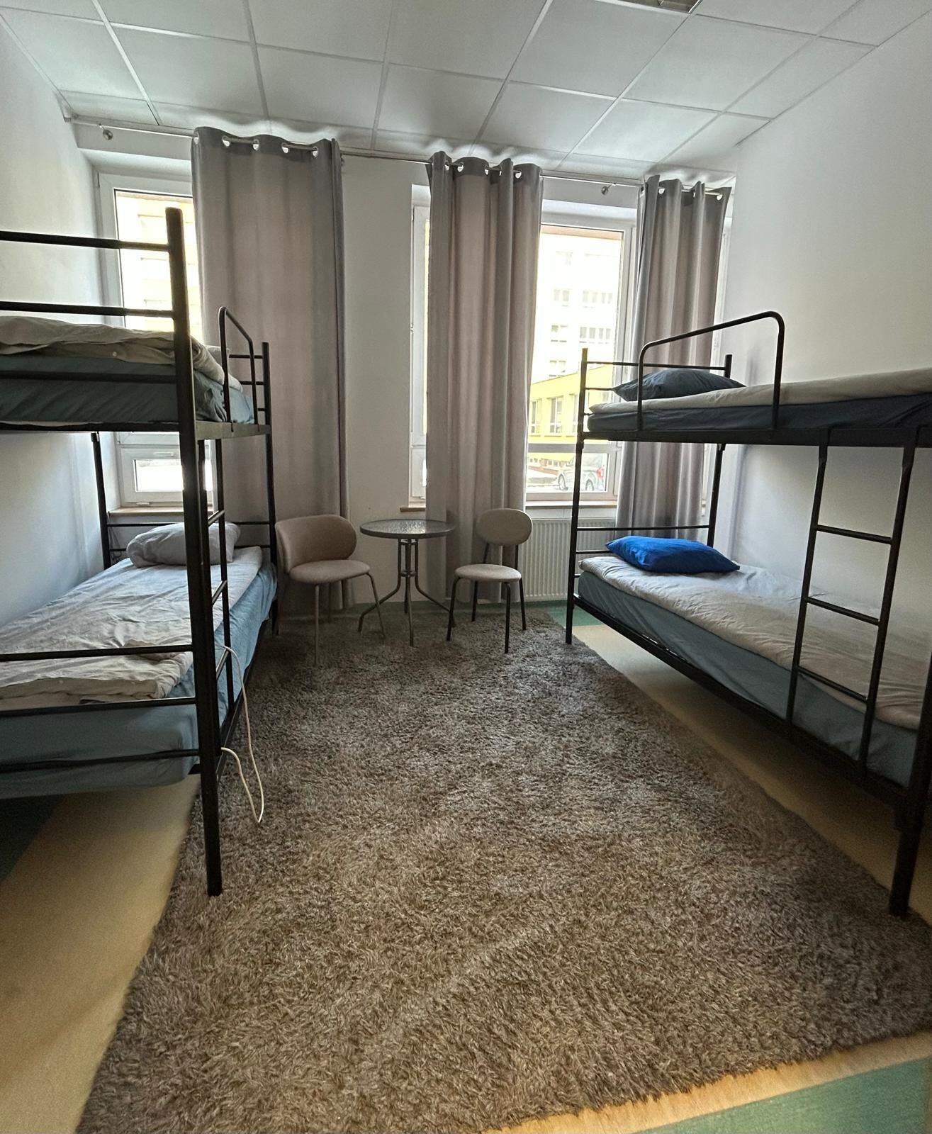 A dormitory room with bunk beds, a small table, two chairs, and a window with curtains.