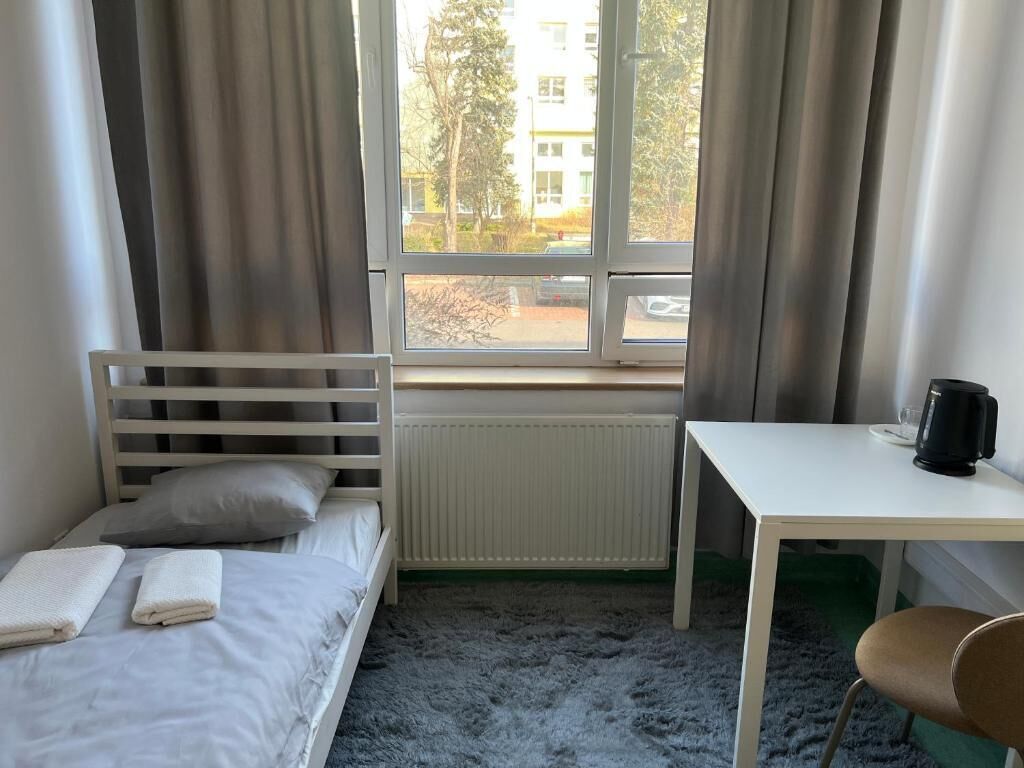 A room with a bed, a table, a chair, a window with curtains, and a radiator.