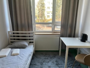 Ein Zimmer mit einem Bett, einem Tisch, einem Stuhl, einem Fenster mit Vorhängen und einer Heizung.