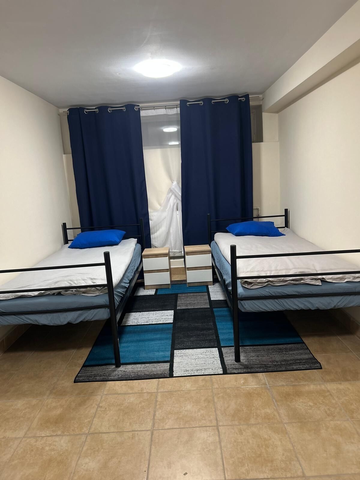 A room with two single beds, a small table, a chair, a window with curtains, and a tiled floor.