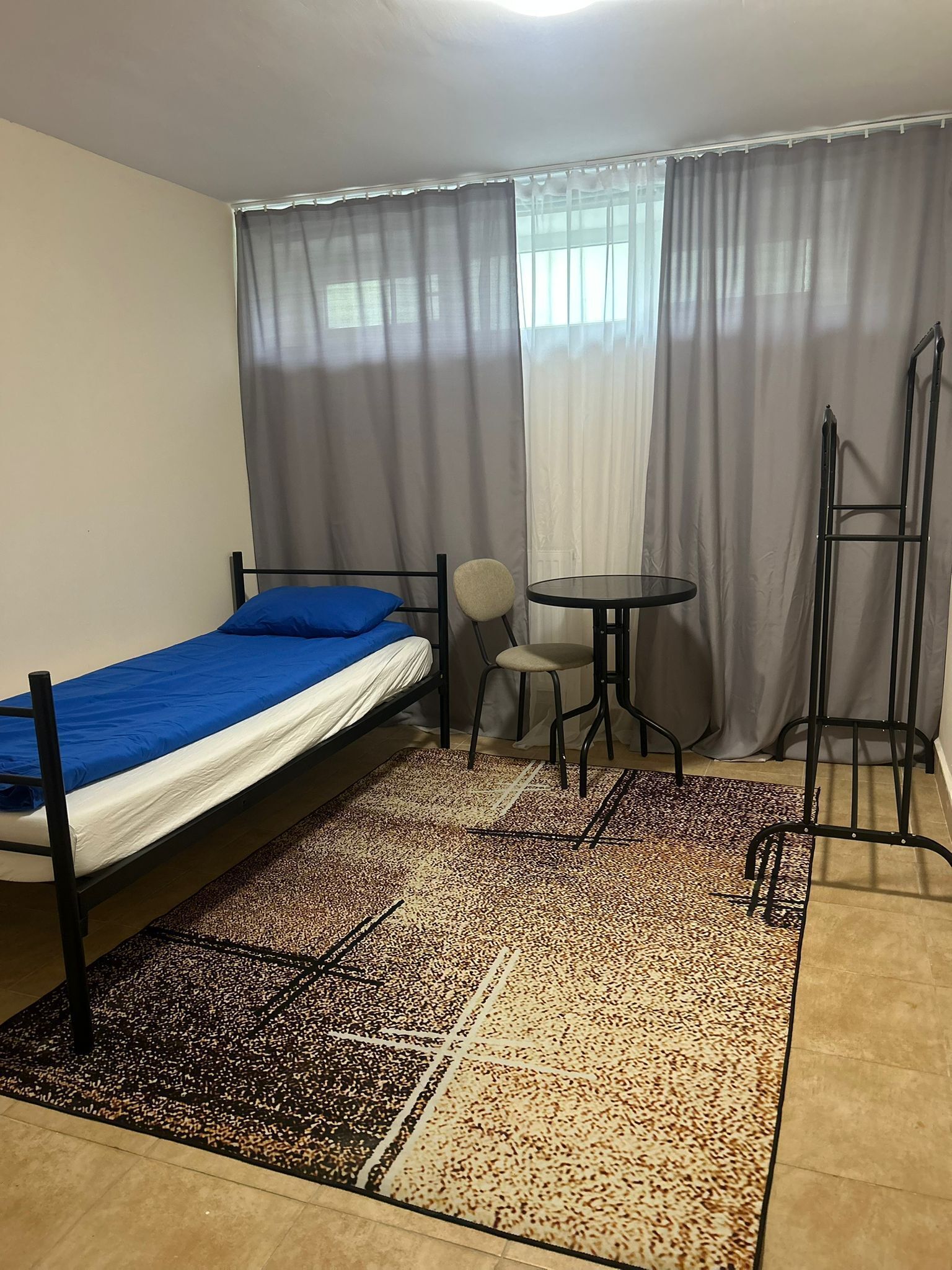 A single bed with a blue cover, a small round table, a chair, and a standing ladder in a room with a patterned rug.