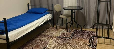 Una cama individual con cubrecolcha azul, una mesita redonda pequeña, una silla y una escalera de pie en una habitación con alfombra estampada.