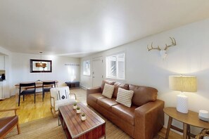 Ein Wohnzimmer mit braunem Ledersofa, einem Couchtisch aus Holz mit Topfpflanzen, einem Essbereich mit Tisch und Stühlen sowie einem an der Wand befestigten Hirschschädel.