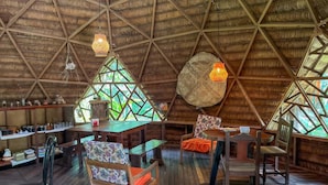 Un coin repas rustique avec des tables et des chaises en bois, un toit de chaume et une grande fenêtre ornée d’un motif géométrique.