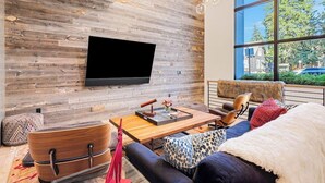 A modern living room with a flat-screen TV mounted on a wooden wall, a sofa, a coffee table, and a large window with a view of the outside. - Luxury townhouse w/ hot tub, great views & incredible location (Jackson)