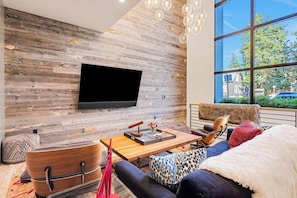 A modern living room with a flat-screen TV mounted on a wooden wall, a sofa, a coffee table, and a large window with a view of the outside. - Luxury townhouse w/ hot tub, great views & incredible location (Jackson)