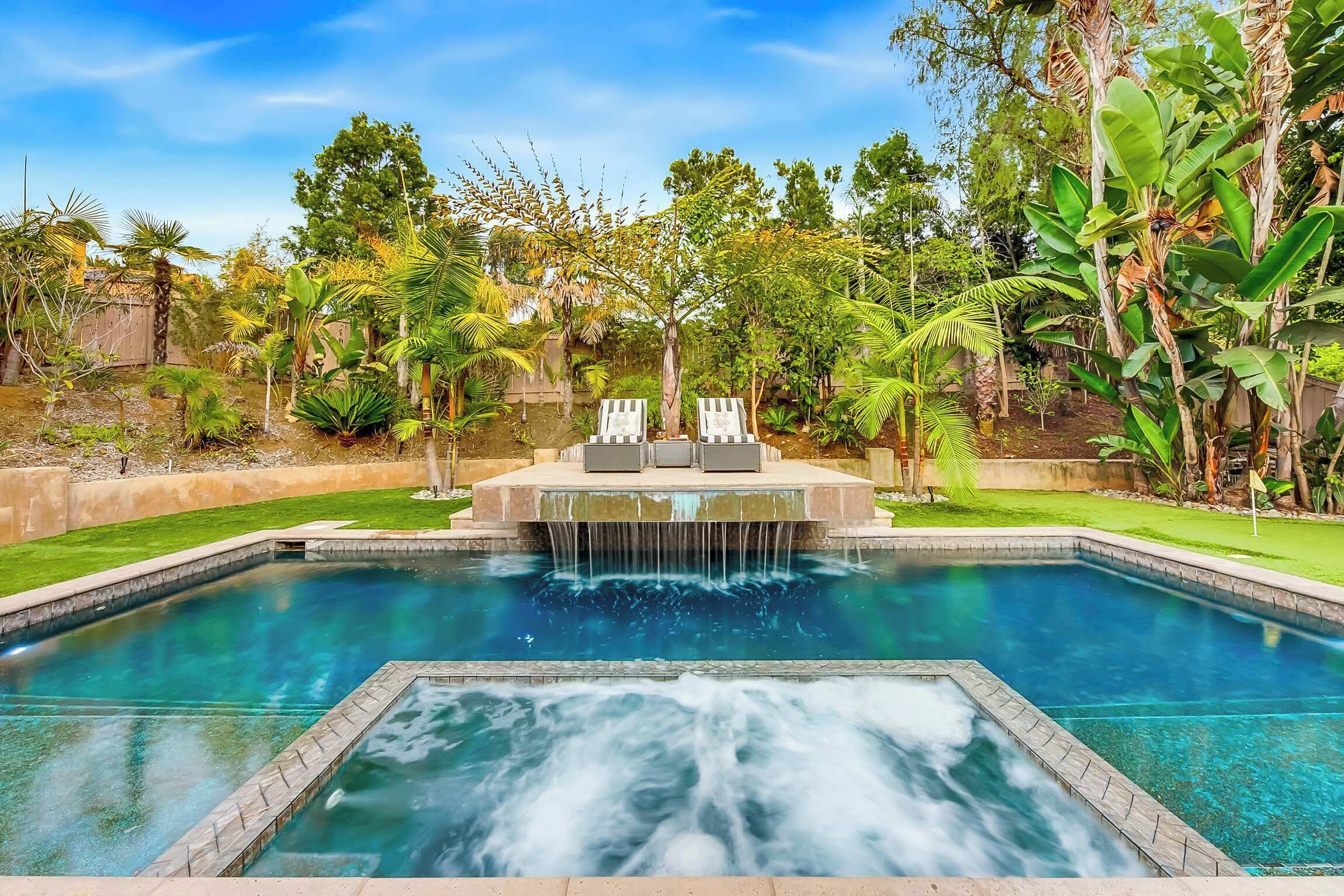 Ein rechteckiger Pool mit Wasserfall, umgeben von üppiger Bepflanzung und einer Holzterrasse mit Liegestühlen.