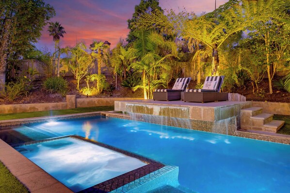 A swimming pool with a waterfall feature, surrounded by lush tropical vegetation and two lounge chairs.