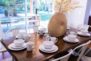 Una mesa de comedor dispuesta con platos, tazones y tazas blancos, un florero con plantas secas y vistas al exterior.
