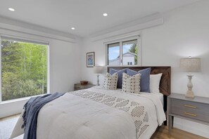 Ein Schlafzimmer mit großem Fenster, einem Bett mit gemustertem Überwurf, einem Nachttisch mit Lampe und Blick auf Bäume.