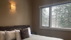 Ein Schlafzimmer mit einem großen Bett, einem Nachttisch, einer Wandleuchte und einem Fenster mit Blick auf Bäume.