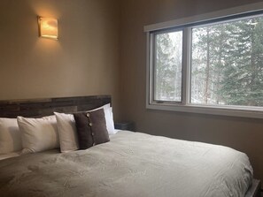 A bedroom with a large bed, a nightstand, a wall-mounted light, and a window with a view of trees. - Riverrfont corner condo with patio, fireplace, electric grill, & washer/dryer (Wilson)