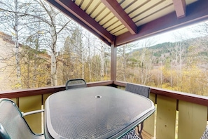 A covered outdoor seating area with a table and chairs, surrounded by a wooden railing, overlooking a forested landscape. - Riverrfont corner condo with patio, fireplace, electric grill, & washer/dryer (Wilson)