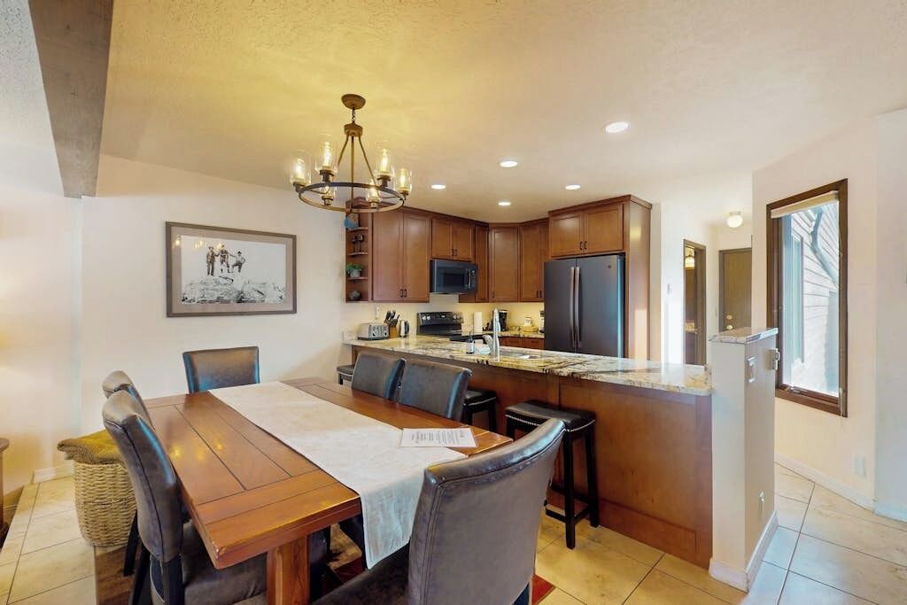 A modern kitchen with a dining area, featuring a chandelier, wooden cabinets, a refrigerator, and a large window.