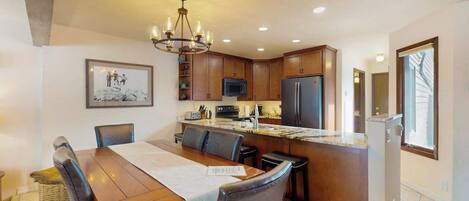 A modern kitchen with a dining area, featuring a chandelier, wooden cabinets, a refrigerator, and a large window.