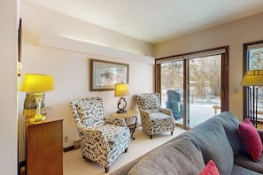 A living room with patterned armchairs, a sofa, a wooden side table, and a lamp.